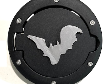 Könnte beinhalten: Schwarzer Tankdeckel-Abdeckung mit einem silbernen Fledermaus-Design. Die Abdeckung ist rund mit einem erhöhten Rand und mehreren Schraubenlöchern am Umfang.