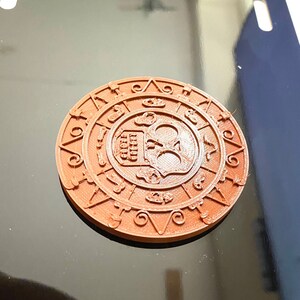 Puede incluir: Un cráneo de color marrón impreso en 3D con un diseño circular a su alrededor. El cráneo tiene un diseño detallado con huesos cruzados y una corona.