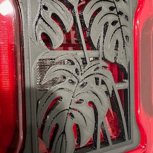 May include: A black plastic cutout of monstera leaves, a tropical plant, is mounted on a red background. The cutout is designed to be placed over a taillight.