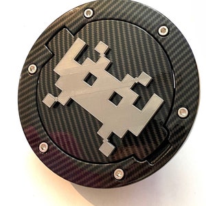 Può includere: Un tappo del serbatoio nero e argento con un motivo in fibra di carbonio e un design di astronave pixelato.