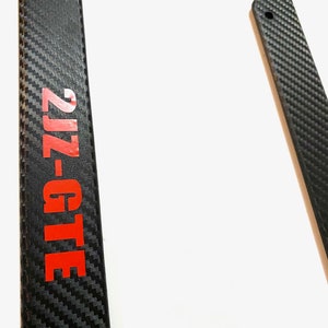 Pode incluir: Moldura de placa de fibra de carbono preta com texto vermelho que diz "2JZ-GTE".