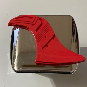 May include: A red 3D printed fin with a textured surface and multiple holes. The fin is sitting on a silver surface.