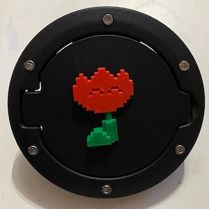 Può includere: Un tappo serbatoio rotondo nero con un design a pixel di un fiore rosso e verde.