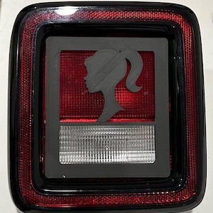 Puede incluir: Un piloto trasero de coche de plástico negro con una lente roja y una luz de marcha atrás blanca. Una silueta de un rostro de mujer está recortada en la lente del piloto trasero.