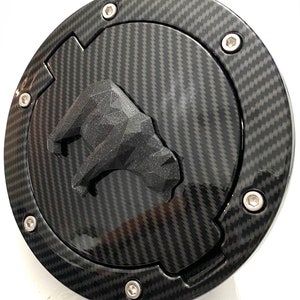 May include: A black carbon fiber gas cap with a 3D bull logo. The cap has a silver ring around the edge and six silver screws.