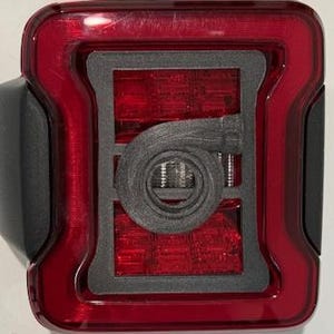 May include: A red and black vehicle tail light with a rectangular shape. The tail light has a black border and a black, circular design in the center. The light is illuminated with red LEDs.