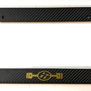 May include: Black carbon fibre license plate frame with a gold logo of a stylised car part and the letters "AE86".