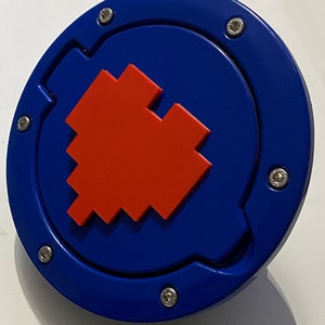 Può includere: Un tappo serbatoio rotondo blu con un design a cuore pixelato rosso. Il tappo ha sei viti argentate attorno al bordo.