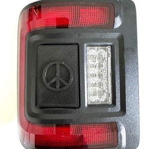 Può includere: Un fanale posteriore nero con una lente rossa e una luce LED bianca. Il fanale posteriore ha un emblema di segno di pace sul lato.