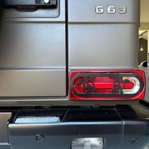Könnte beinhalten: Ein grauer SUV mit schwarzer Stoßstange hinten und einem roten Rücklicht mit einem schwarzen Schildkröten-Design. Das Fahrzeug hat ein G63-Emblem hinten.