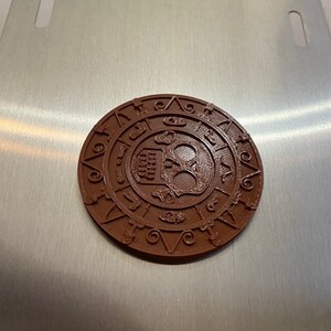 Puede incluir: Una placa circular de metal marrón con un diseño de calavera y huesos cruzados y un círculo de números y símbolos alrededor del exterior.