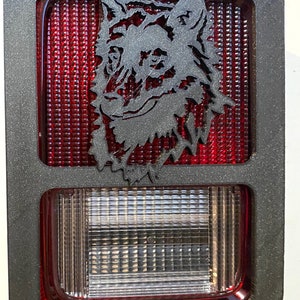 Puede incluir: Marco de metal negro con un recorte de silueta de lobo sobre una luz trasera roja y transparente. La luz trasera tiene un patrón de rejilla.