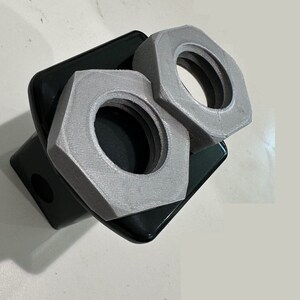 May include: Two silver metal nuts, one on top of the other, are attached to a black metal object. The nuts are hexagonal in shape and have a smooth, shiny surface.