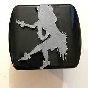 May include: A gray plastic hula girl silhouette on a black square background.