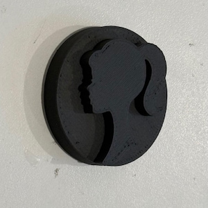 May include: A black silhouette of a woman's face with a ponytail, printed on a round, black, 3D-printed object.
