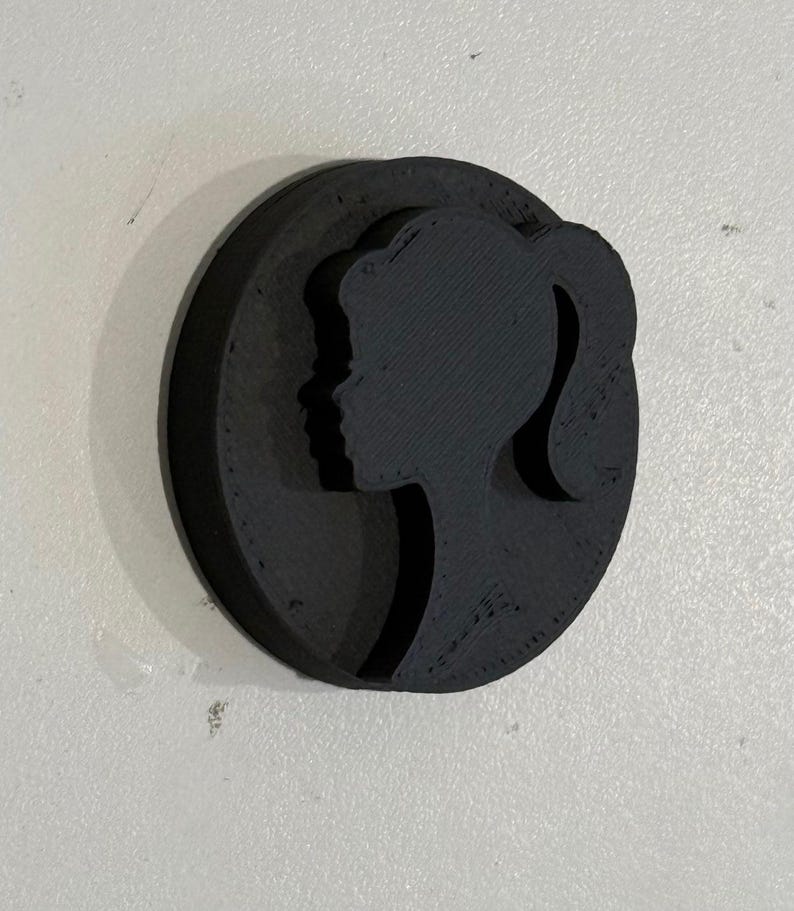 May include: A black silhouette of a woman's face with a ponytail, printed on a round, black, 3D-printed object.