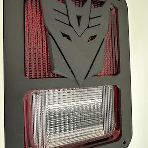 May include: Black plastic tail light cover with a red lens and a cutout in the shape of the Transformers logo. The logo is black and is in the center of the cover.