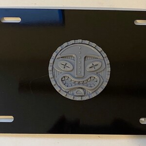 May include: A black license plate with a gray, raised, tiki mask design in the center.