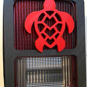 Peut inclure: Un cadre en plastique noir avec un feu arrière rouge et une découpe de tortue rouge. Le feu arrière a un objectif blanc en dessous.
