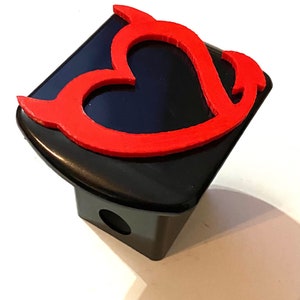 Puede incluir: Una tapa de enganche de metal negro con un diseño de corazón rojo con cuernos en la parte superior.