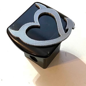 Puede incluir: Una tapa de enganche de metal negro con un diseño de corazón plateado. El corazón tiene un diseño estilizado y fluido.