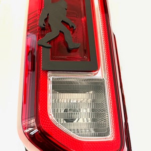May include: Red taillight with a black silhouette of a Bigfoot walking inside a black frame.