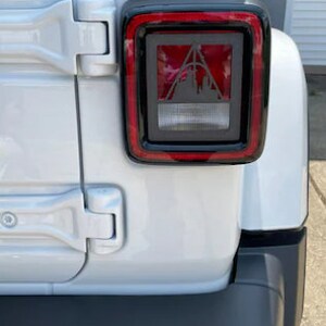 Puede incluir: Jeep Wrangler blanco con un parachoques trasero negro y una luz trasera negra con una lente roja. La luz trasera tiene una calcomanía negra con un gráfico de un castillo.