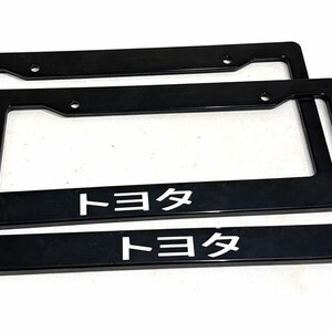 May include: Two black license plate frames with the Japanese text "Toyota" in white. The frames are rectangular with rounded corners and mounting holes. The frames are made of a glossy material.