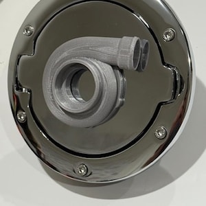 May include: A chrome-finished fuel door with a gray, 3D-printed turbocharger design. The door is circular with a recessed center and is secured by six small, silver-colored screws. The turbocharger is a detailed model of the engine component.