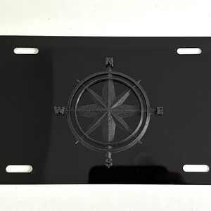 May include: Black license plate with a raised compass design. The compass has the cardinal directions N, S, E, and W.