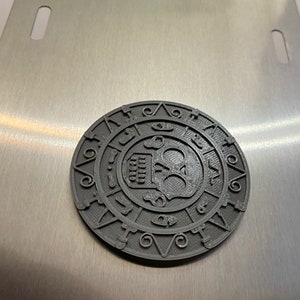 Puede incluir: Un posavasos circular de metal gris impreso en 3D con un diseño de calavera en el centro. La calavera está rodeada por un círculo de números y símbolos.