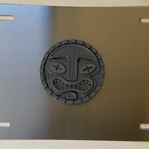 May include: A silver metal license plate frame with a gray plastic tiki mask design in the center.