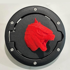 Puede incluir: Una tapa de gasolina negra con una cabeza de Godzilla roja impresa en 3D en la parte superior.