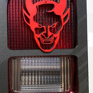 Op de afbeelding: Een zwart kunststof frame met een rood achterlicht en een rode duivelskop-uitsparing. Het achterlicht heeft een witte lens met een rasterpatroon.