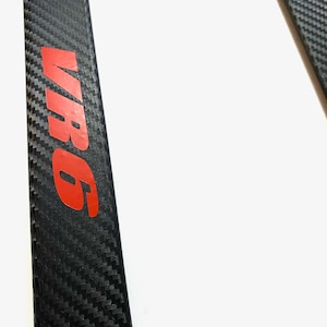 May include: A black carbon fibre trim piece with the word "VR6" in red lettering.