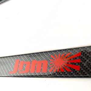 Könnte beinhalten: Ein schwarzer Kennzeichenrahmen aus Kohlefaser mit einem roten "JDM"-Logo und einer roten aufgehenden Sonne.
