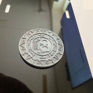 Puede incluir: Un diseño de calavera y huesos cruzados de metal gris sobre un fondo negro. La calavera está rodeada por un círculo de símbolos y números.