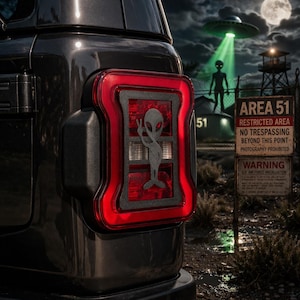 May include: Black vehicle tail light with a red border and a gray alien graphic giving a peace sign. The background shows an Area 51 sign, a UFO, and an alien figure under a night sky.