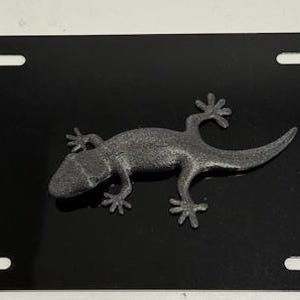 Puede incluir: Placa de matrícula negra con un emblema de lagarto gris detallado. La placa tiene cuatro ranuras rectangulares para el montaje. El lagarto está en posición de reptar, con las patas y la cola extendidas.