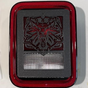 Puede incluir: Luz trasera rectangular roja y negra con un detallado emblema de cabeza de lobo. El emblema está centrado en un fondo gris oscuro, con una sección de luz clara debajo. El marco exterior es rojo brillante.