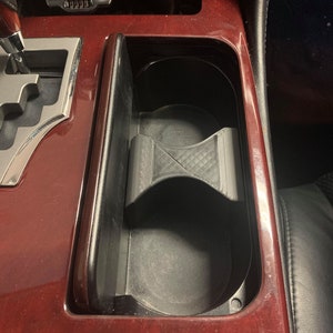 May include: A close-up of a car's interior with a black phone holder in a cup holder. The cup holder is located in the center console of the car. The console is made of wood and has a red finish.