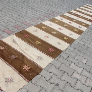 May include: A long, narrow rug with a striped pattern in shades of brown, beige, and white. The rug has a fringe on both ends and features a repeating star design.