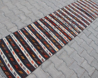 May include: A long, narrow rug with a striped pattern in shades of black, white, red, orange, and yellow. The rug has a traditional, handwoven look.