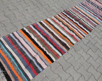 Può includere: Un tappeto lungo e stretto a strisce multicolori con bordi sfrangiati. Il tappeto è realizzato in tessuto tessuto e presenta una varietà di colori, tra cui arancione, blu, rosso e nero.