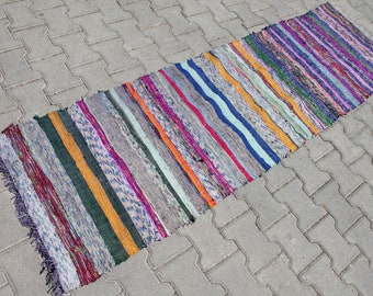 Può includere: Un tappeto a strisce colorato con una varietà di colori, tra cui rosso, arancione, giallo, verde, blu, viola e grigio. Il tappeto è realizzato in tessuto tessuto e ha un bordo con frange.