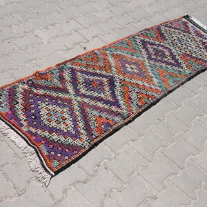 May include: A long, narrow rug with a geometric pattern in shades of orange, purple, green, and white. The rug has a fringe on both ends.