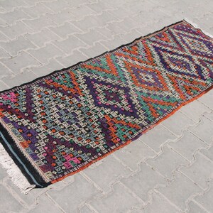 May include: A colourful, handwoven rug with a geometric pattern in shades of orange, purple, green, and black. The rug has a fringe on both ends.