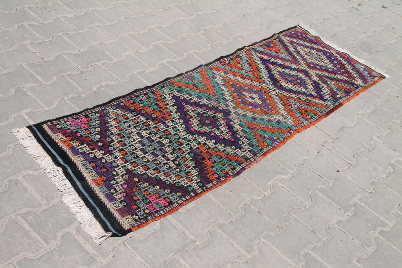 May include: A colourful, handwoven rug with a geometric pattern in shades of orange, purple, green, and black. The rug has a fringe on both ends.