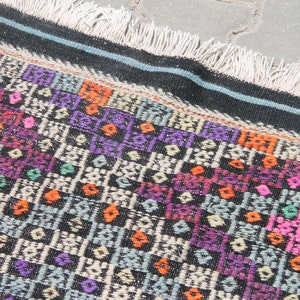May include: A colourful woven rug with a geometric pattern in shades of pink, purple, orange, and green on a black background. The rug has a fringe border.