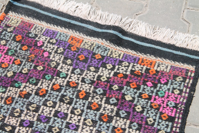 May include: A colourful woven rug with a geometric pattern in shades of pink, purple, orange, and green on a black background. The rug has a fringe border.